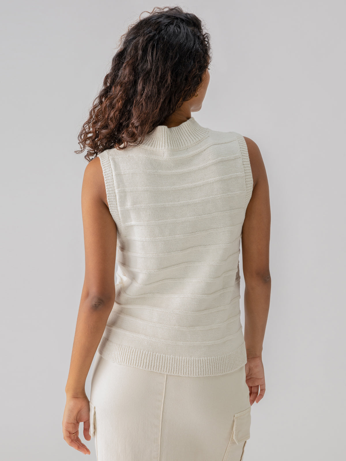 A person with curly hair is seen from behind, wearing Sanctuary Clothing's Life Is Easy Shell Chalk, a sleeveless, white, ribbed sweater paired with a matching skirt. The background is plain light gray.