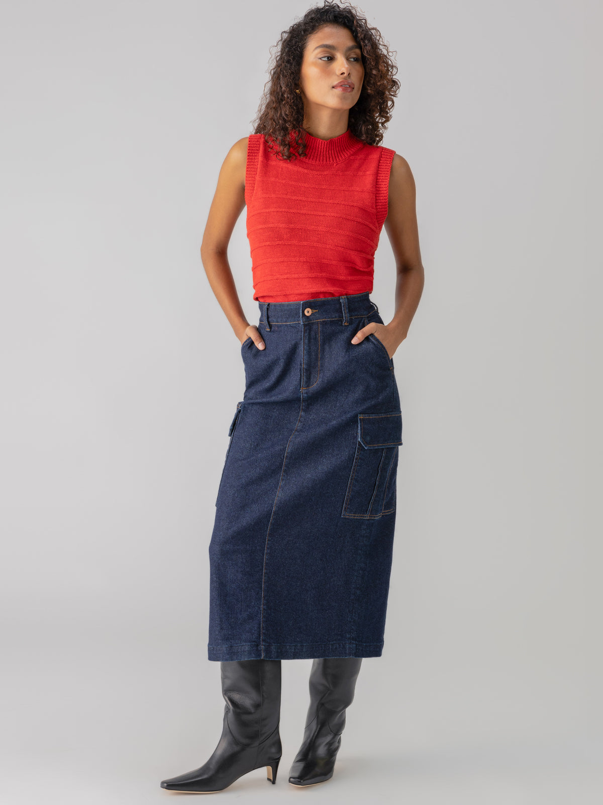 A person with curly hair stands confidently with hands in pockets, wearing the Life Is Easy Shell Cherry Red top from Sanctuary Clothing and a high-waisted, knee-length dark blue denim skirt with a front pocket. They also wear knee-high black boots. They are set against a plain grey background.