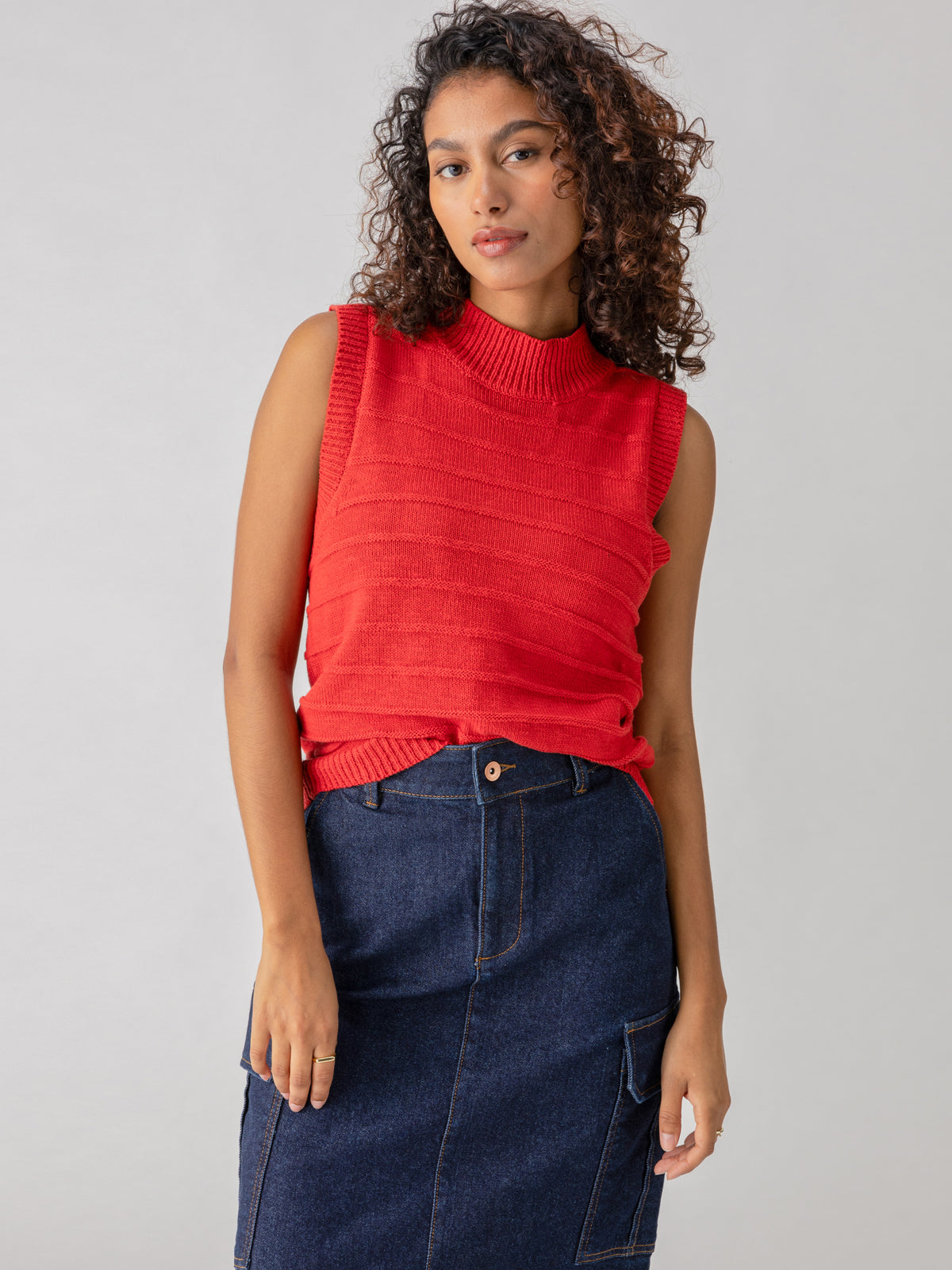 A person with curly hair is wearing the Sanctuary Clothing Life Is Easy Shell Cherry Red and a high-waisted, dark blue denim skirt. They are standing against a plain gray background, looking confidently at the camera with a relaxed expression.