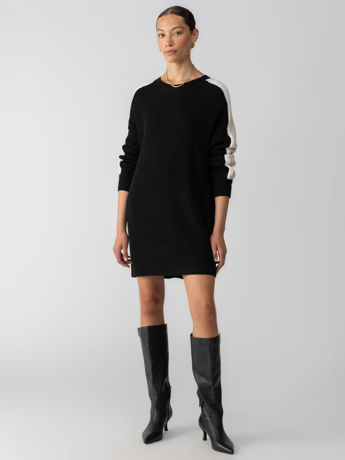 A person stands against a plain background, wearing the Speedway Sweater Dress in Black Milk Toasted Almond from Sanctuary Clothing. They have their hair pulled back and are dressed in black knee-high boots. The sweater dress has a relaxed fit and ends mid-thigh, featuring white stripes down the sleeves.