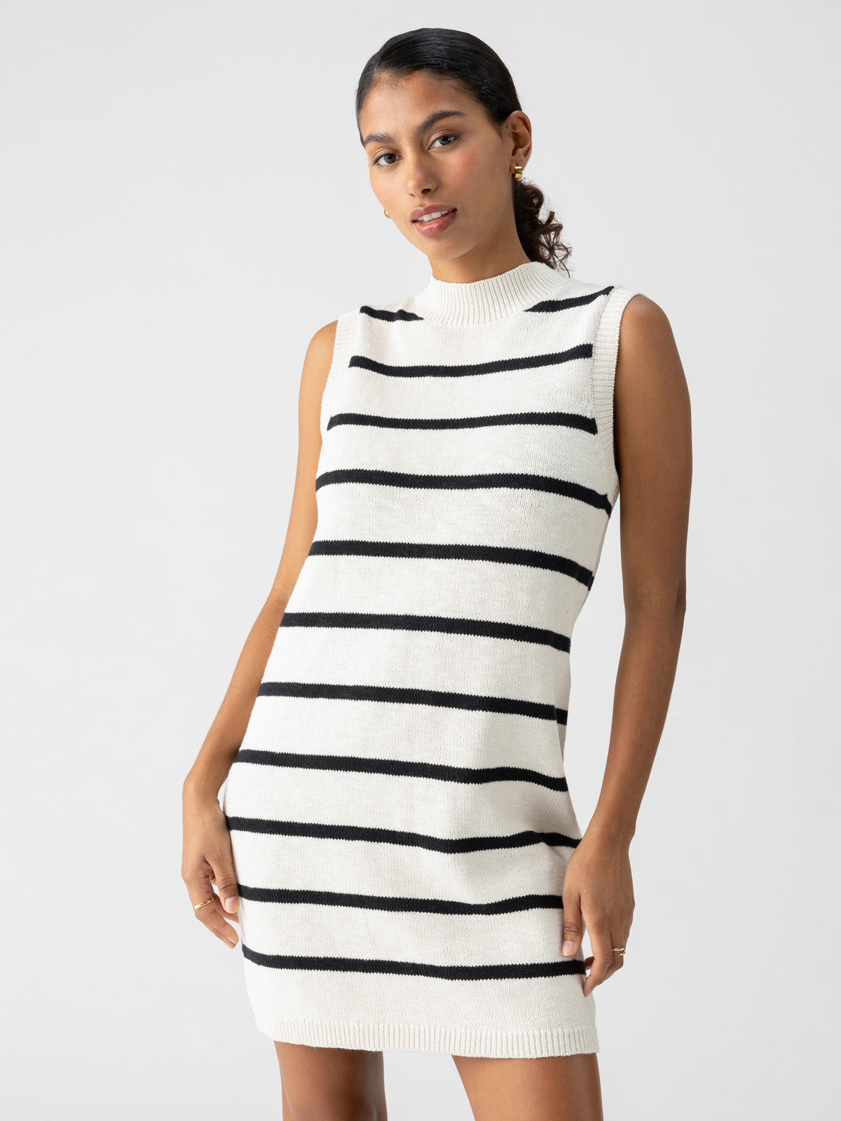 A person wearing the Sanctuary Clothing "Life Is Easy Swtr Dress Chalk Black Stripe," a sleeveless, high-neck, white sweater dress with horizontal black stripes, stands confidently with hands resting on their hips. The plain white background highlights the simplicity and elegance of the outfit.