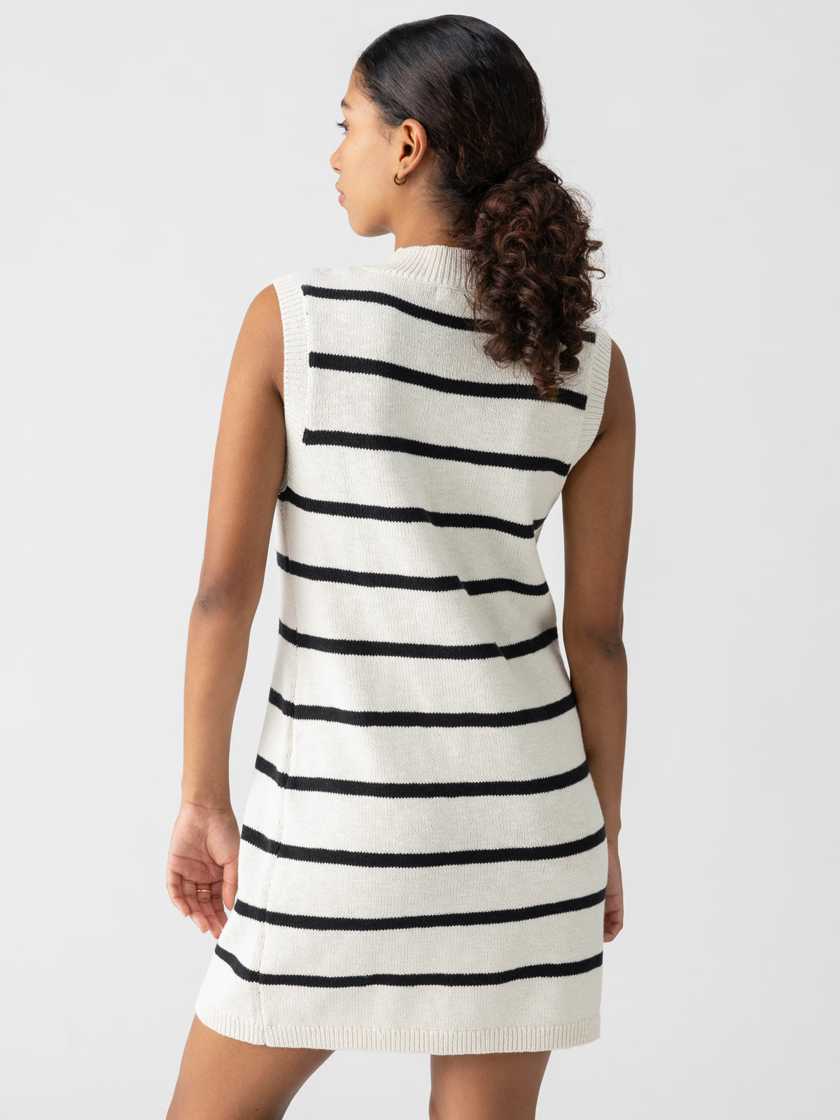 A woman, with her dark hair pulled back into a low ponytail, stands with her back to the camera. She is donning the "Life Is Easy Swtr Dress Chalk Black Stripe" by Sanctuary Clothing—a sleeveless, knee-length dress adorned with black horizontal stripes against a white backdrop. The background is plain and white.