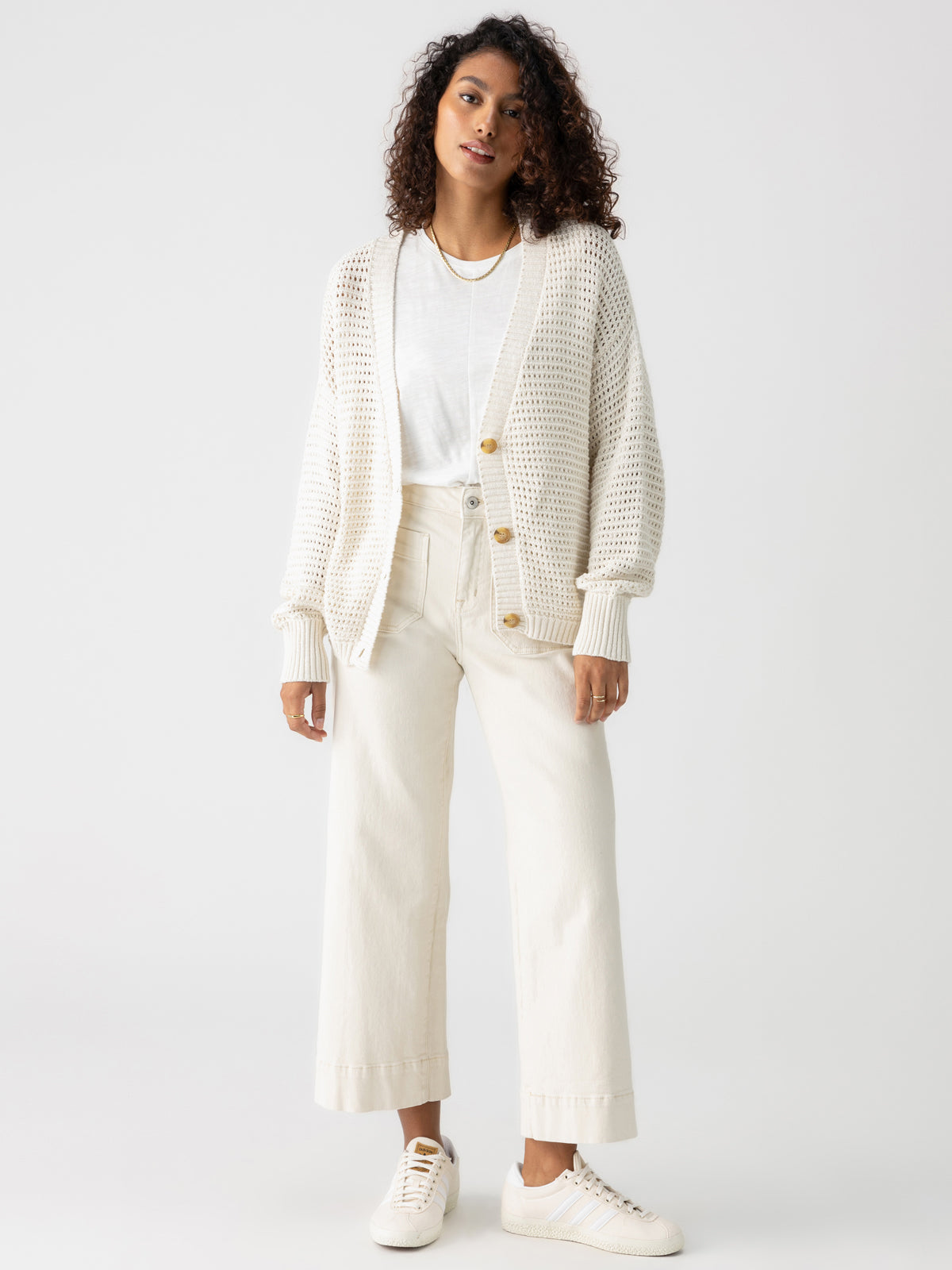 A person with curly hair stands wearing the Sanctuary Clothing Falling For Fall Cardi Chalk over a white t-shirt, beige wide-legged pants, and white sneakers. The background is plain and white, highlighting the casual, monochromatic outfit.