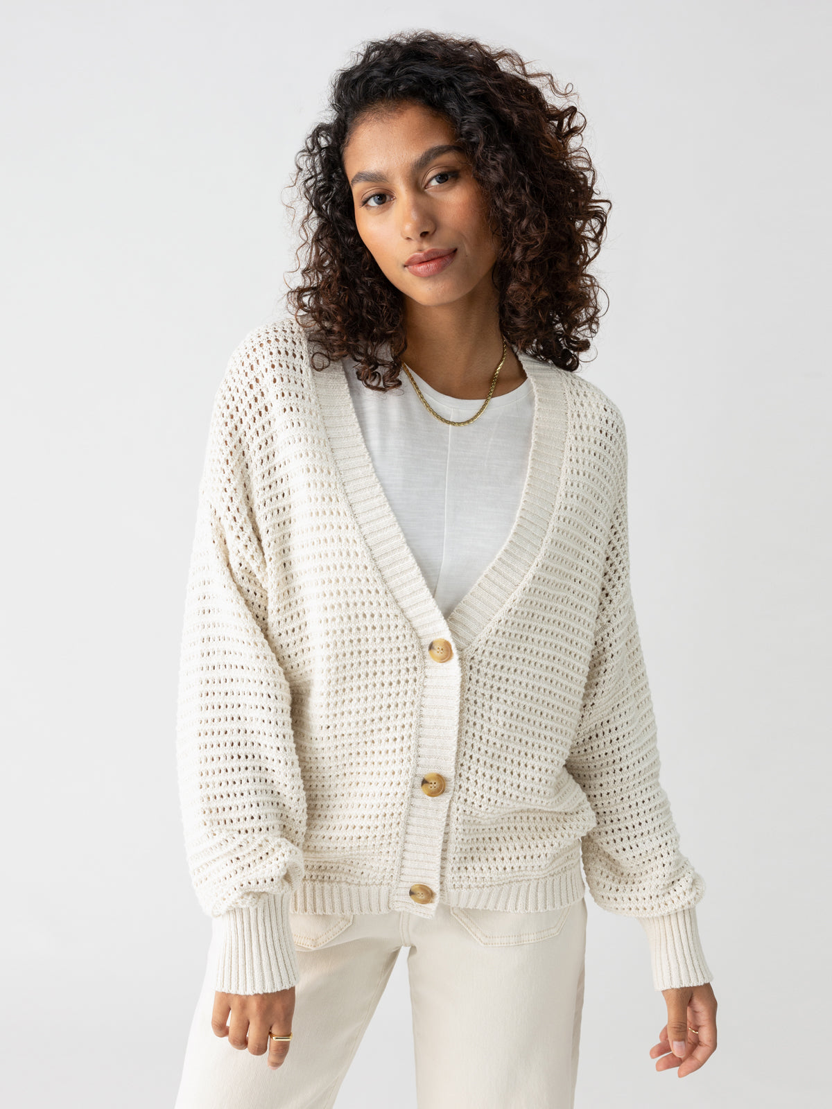 A person with curly hair is wearing the "Falling For Fall Cardi" in Chalk by Sanctuary Clothing over a white t-shirt. They are looking calmly at the camera against a plain gray background.