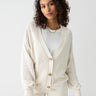 A person with curly hair is wearing the "Falling For Fall Cardi" in Chalk by Sanctuary Clothing over a white t-shirt. They are looking calmly at the camera against a plain gray background.