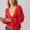 A person with shoulder-length hair wears the vibrant Sanctuary Clothing Falling For Fall Cardi in Cherry Red over a white top and blue pants. They have one hand holding a corner of the cardigan and the other hand is partially visible. They also sport a gold necklace and rings while gazing directly at the camera.