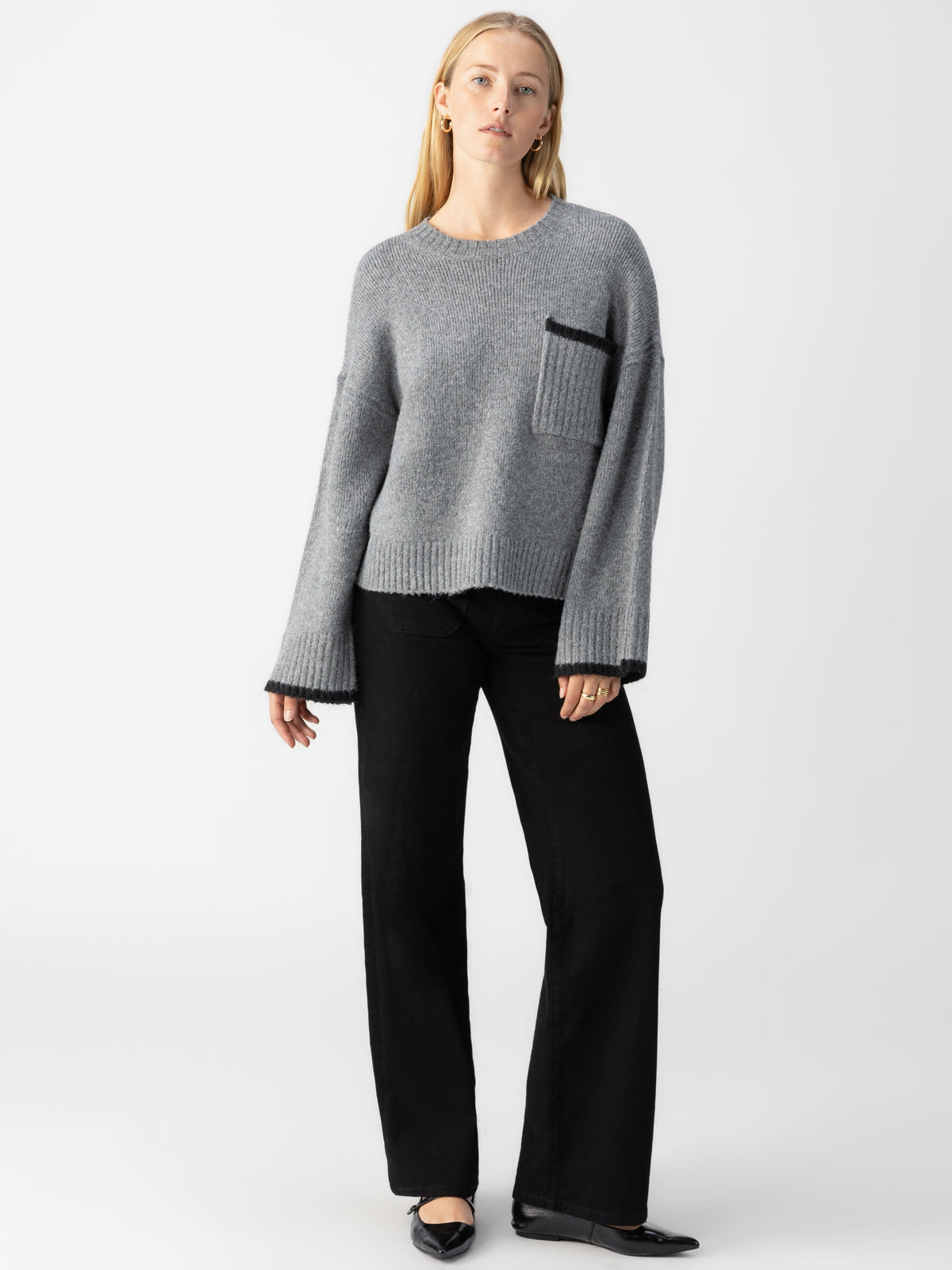 A person stands against a plain white background, wearing the Uptown Girl Sweater in Heather Ash by Sanctuary Clothing along with black trousers. Their hands are at their sides, and they are wearing black shoes. They have long, light-colored hair and a neutral expression.