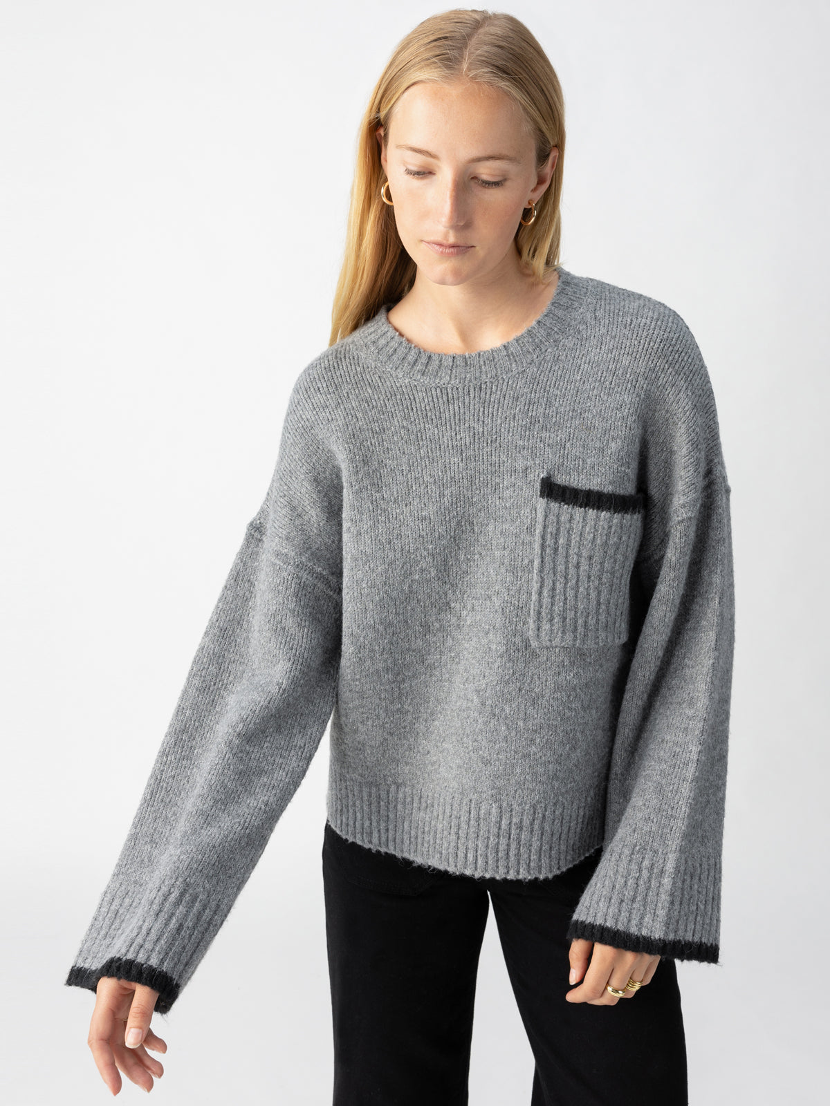 A person with long, straight hair wearing the Uptown Girl Sweater Heather Ash by Sanctuary Clothing, which features a loose fit, a breast pocket, and wide sleeves. They are looking down and slightly to the side while standing in front of a plain white background. The individual is also dressed in black pants.