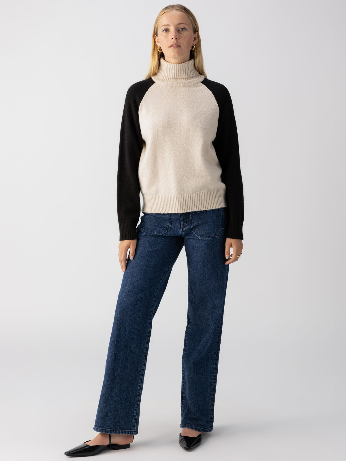 A person with long hair stands against a plain background, confidently donning the Cozy Day Sweater in Toasted Almond Black by Sanctuary Clothing. They match it with blue jeans and black pointed shoes. Sporting a neutral expression, they pose casually with their arms relaxed by their sides.