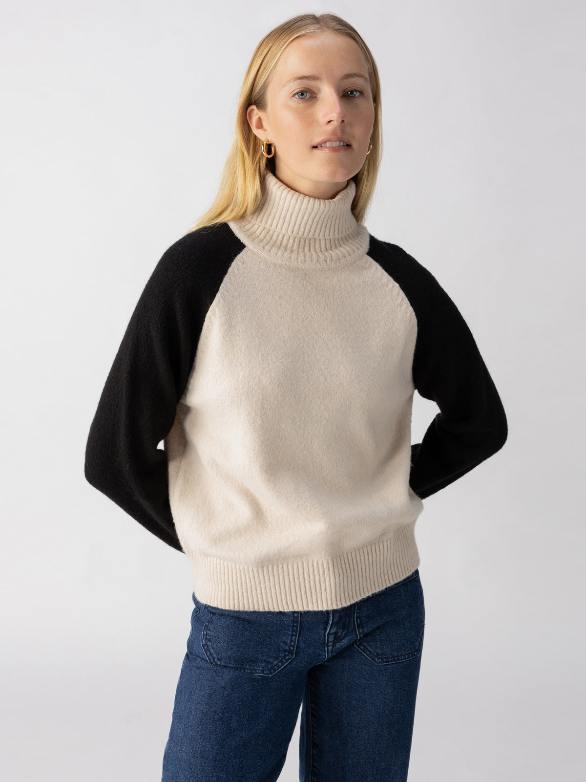 A person with long blonde hair is wearing a cream-colored Cozy Day Sweater in the Toasted Almond Black style by Sanctuary Clothing, paired with blue jeans. They have a neutral expression and are standing in front of a plain, light gray background. Their hands are behind their back.