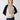 A person with long blonde hair is wearing a cream-colored Cozy Day Sweater in the Toasted Almond Black style by Sanctuary Clothing, paired with blue jeans. They have a neutral expression and are standing in front of a plain, light gray background. Their hands are behind their back.