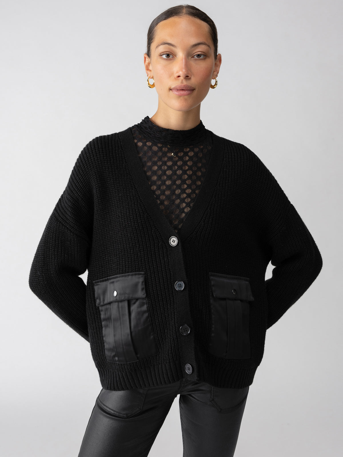 A person with hair pulled back and gold earrings wears the Trend Setter Cardi Black from Sanctuary Clothing, which features buttons and front pockets, layered over a black polka-dot top. They stand against a plain light gray background with hands resting on their hips.