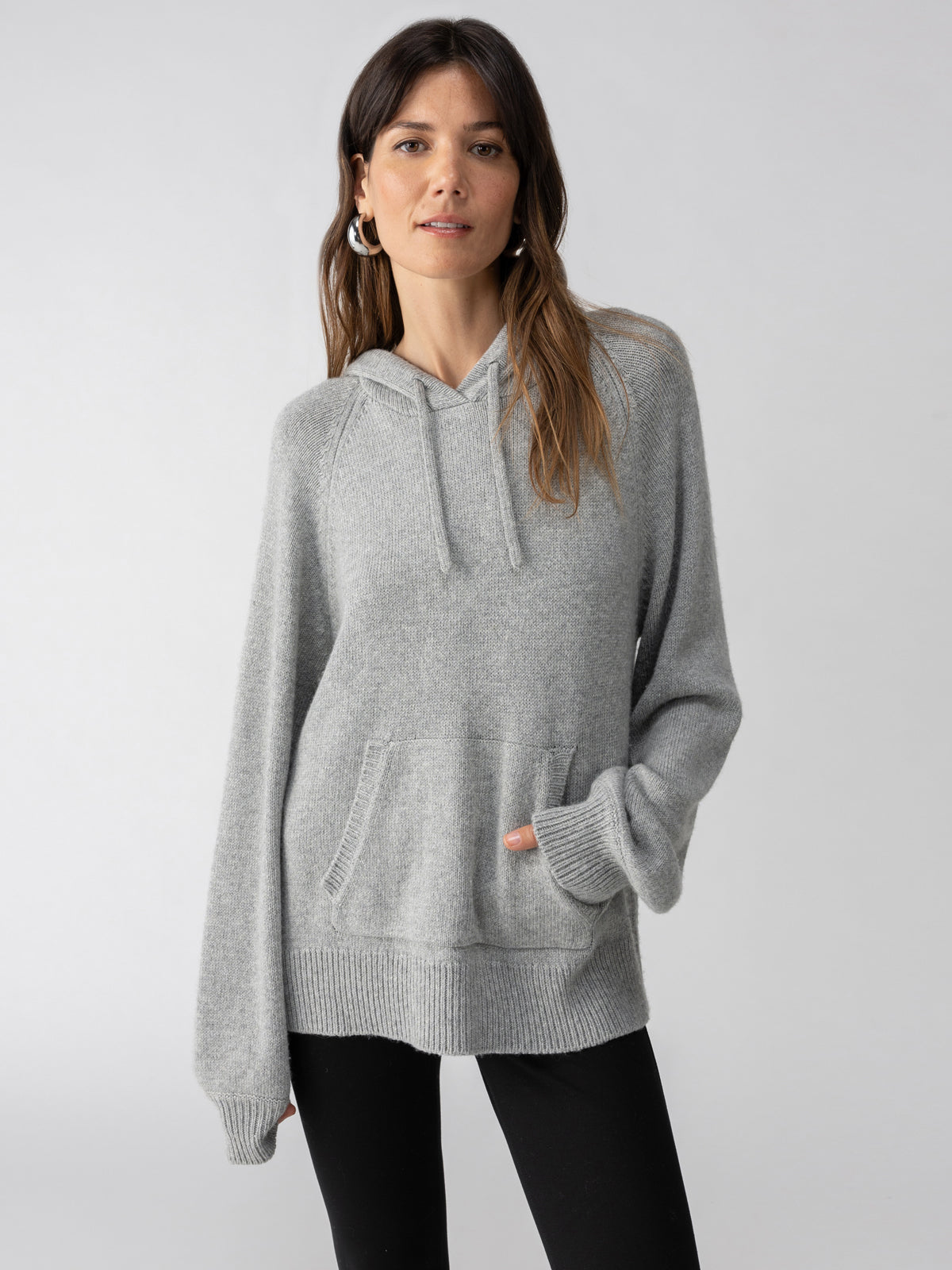 A woman with long hair is wearing the Sanctuary Clothing Elevated Layering Hoodie in Heather Grey and black pants. She stands against a plain, light background, one hand in her pocket, looking at the camera with a neutral expression.