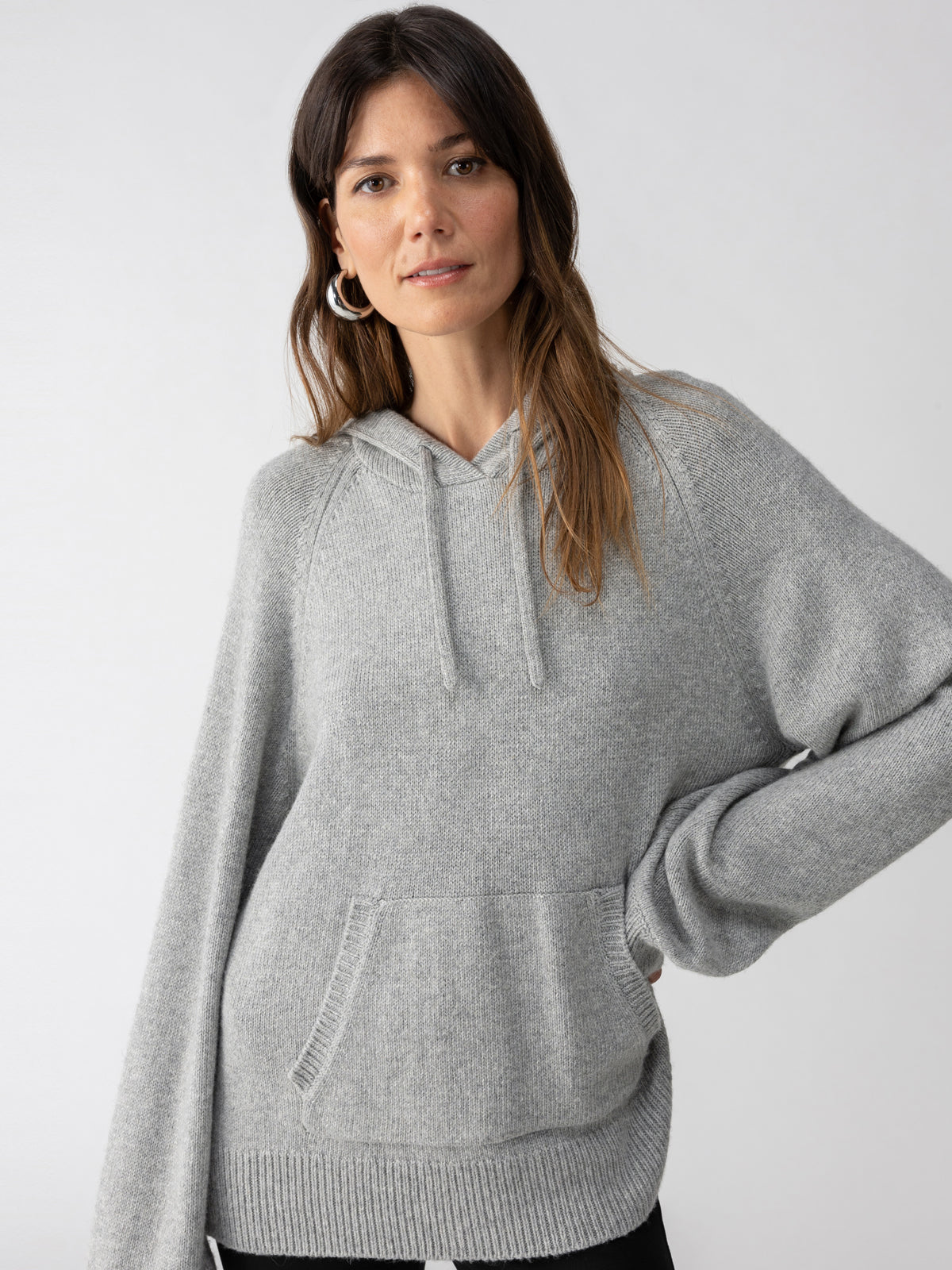 A woman with long brown hair is wearing the Sanctuary Clothing Elevated Layering Hoodie in Heather Grey and black pants. She stands against a plain white background, looking directly at the camera with a relaxed expression and her right hand on her hip.