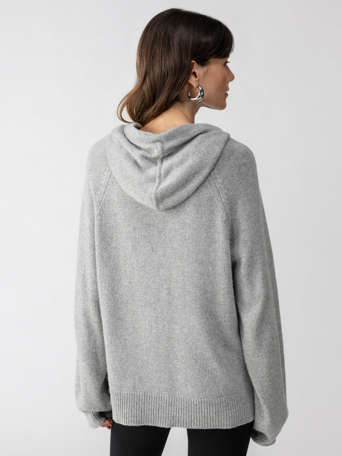 A person with long hair is facing away, wearing a loose-fitting Elevated Layering Hoodie in Heather Grey by Sanctuary Clothing. They have round hoop earrings, and the background is plain and off-white.