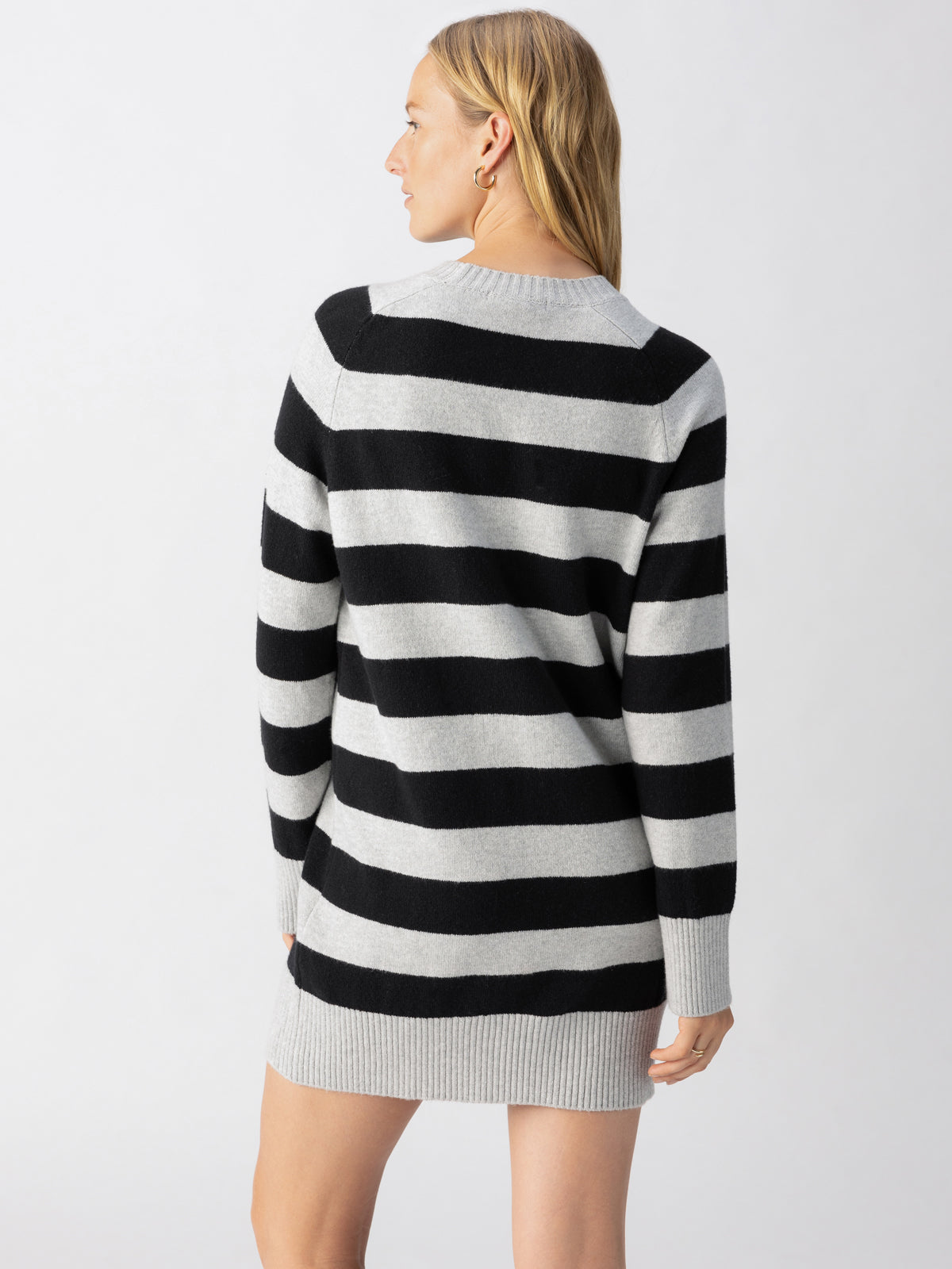 A person with long blonde hair is shown from the back, wearing the Sanctuary Clothing So Soft Sweater Dress Heather Grey Black Stripe, a long-sleeved, knee-length sweater dress featuring wide black and gray horizontal stripes. The background is plain and white.