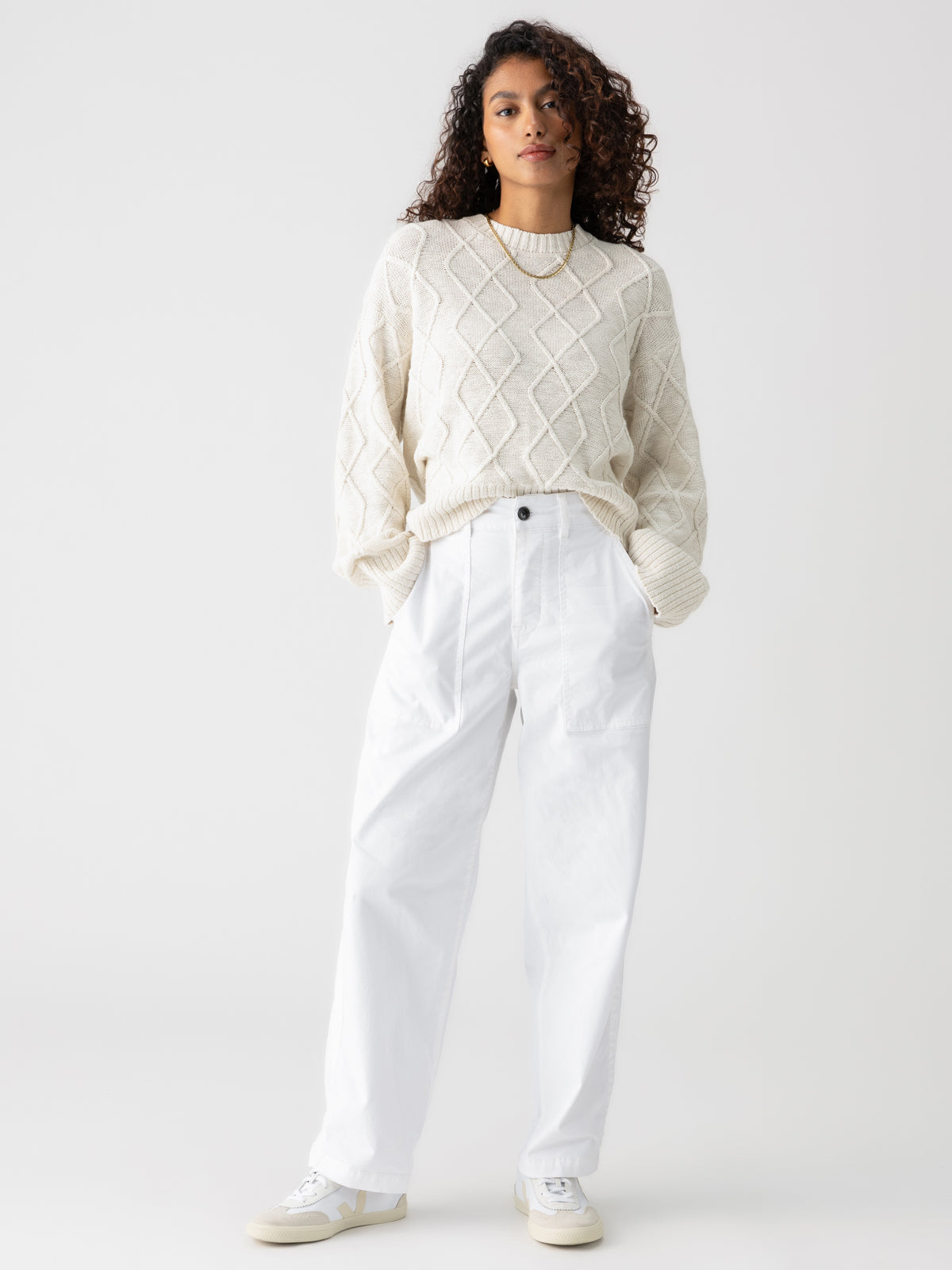 A person with curly hair stands against a light grey background. They are wearing the Coastal Cable Crewneck Chalk by Sanctuary Clothing and white, high-waisted, wide-legged trousers. They have their hands in their pockets and are wearing white sneakers.