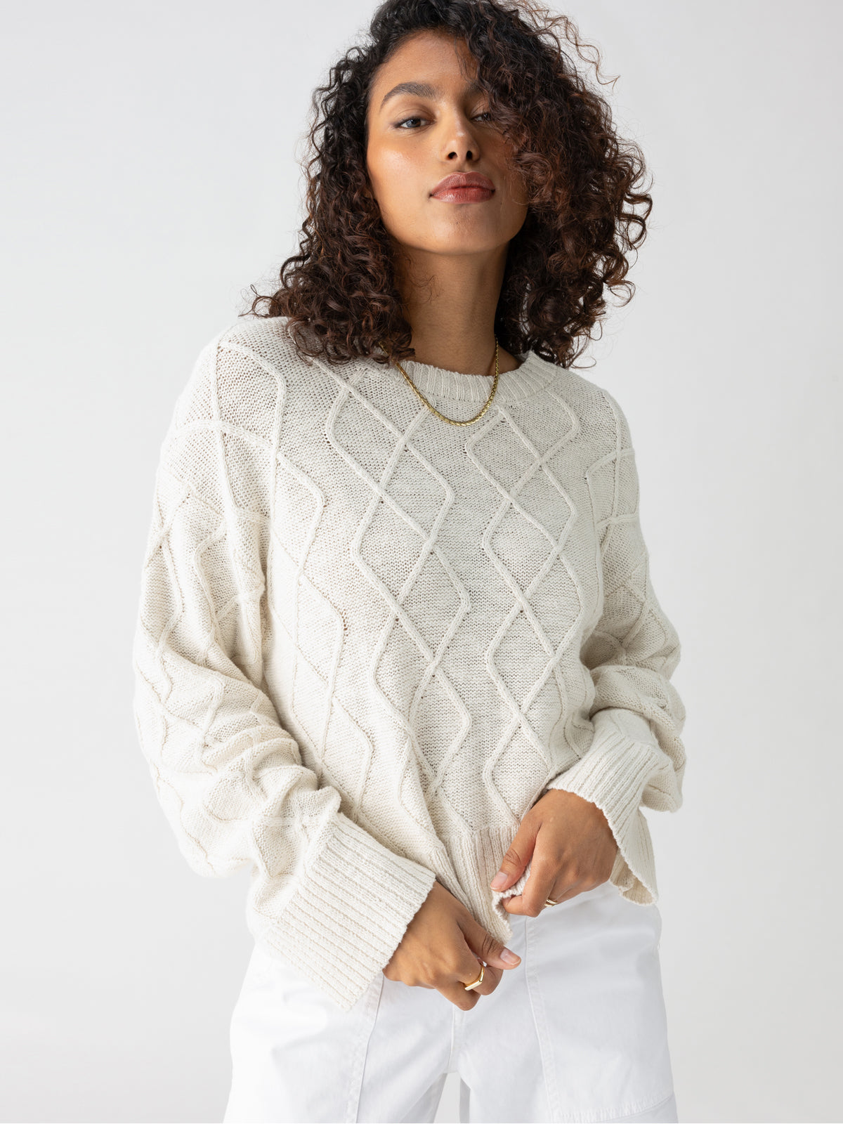 A person with curly hair is wearing the Coastal Cable Crewneck Chalk from Sanctuary Clothing, a cream-colored, textured knit sweater featuring a diamond pattern. They pair it with white pants and accessorize with a simple gold necklace. The individual stands against a plain, white background with a subtle expression.