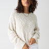 A person with curly hair is wearing the Coastal Cable Crewneck Chalk from Sanctuary Clothing, a cream-colored, textured knit sweater featuring a diamond pattern. They pair it with white pants and accessorize with a simple gold necklace. The individual stands against a plain, white background with a subtle expression.