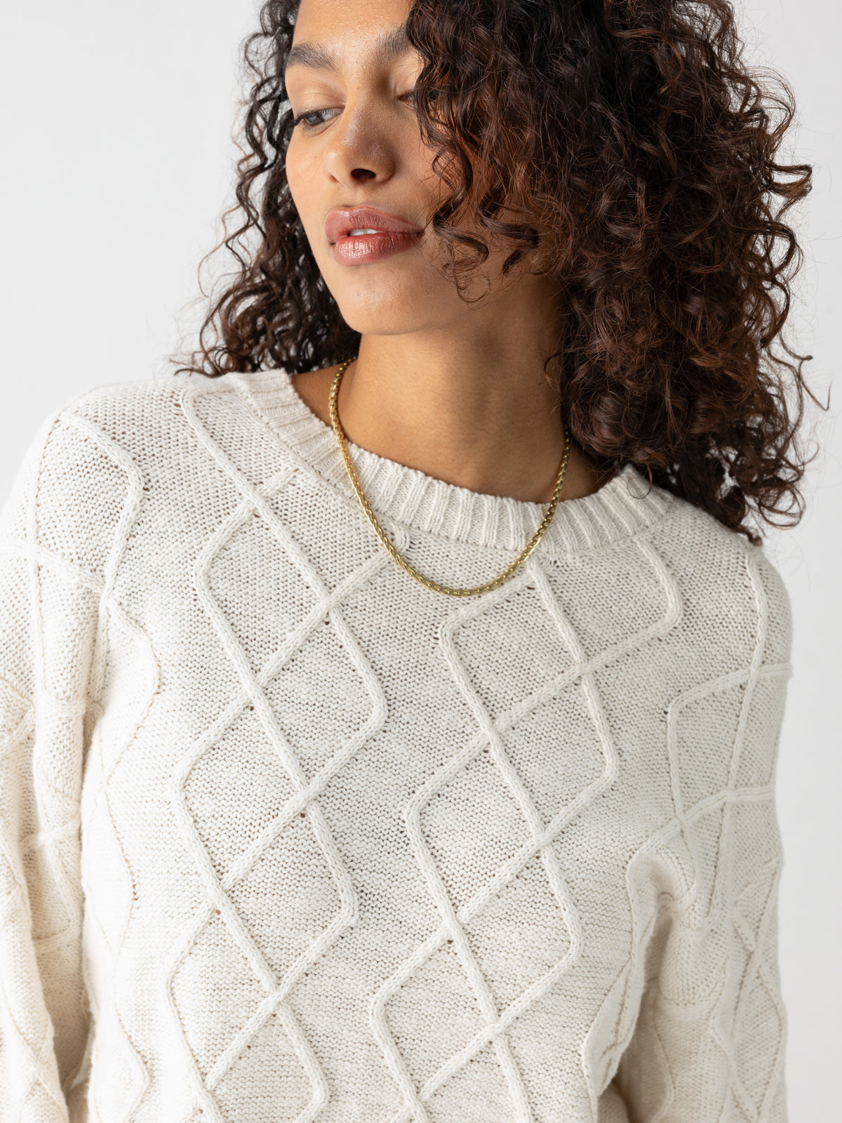 A person with curly brown hair is wearing the Coastal Cable Crewneck Chalk sweater from Sanctuary Clothing, featuring a textured diamond pattern. They accessorize with a gold necklace and pose slightly downwards and sideways against a plain light background.