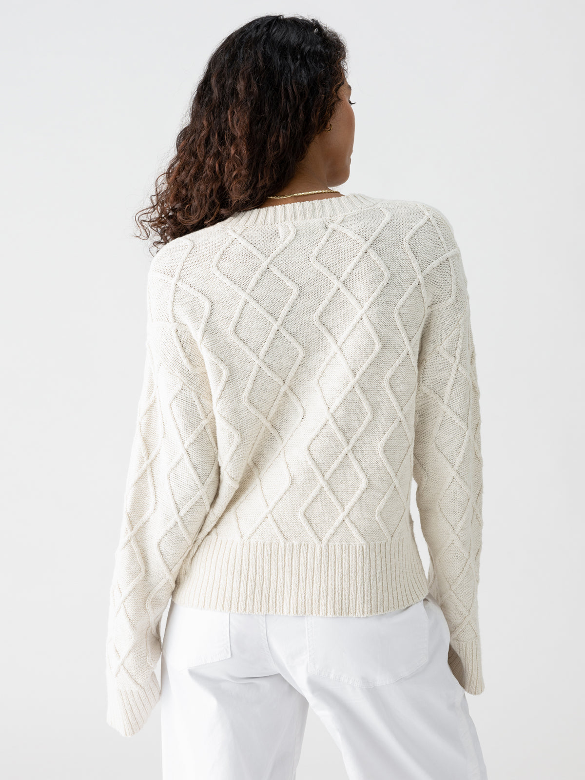 A person with long, curly hair is standing with their back to the camera, wearing a cream-colored Coastal Cable Crewneck Chalk sweater from Sanctuary Clothing and white pants. The background is plain and light-colored.