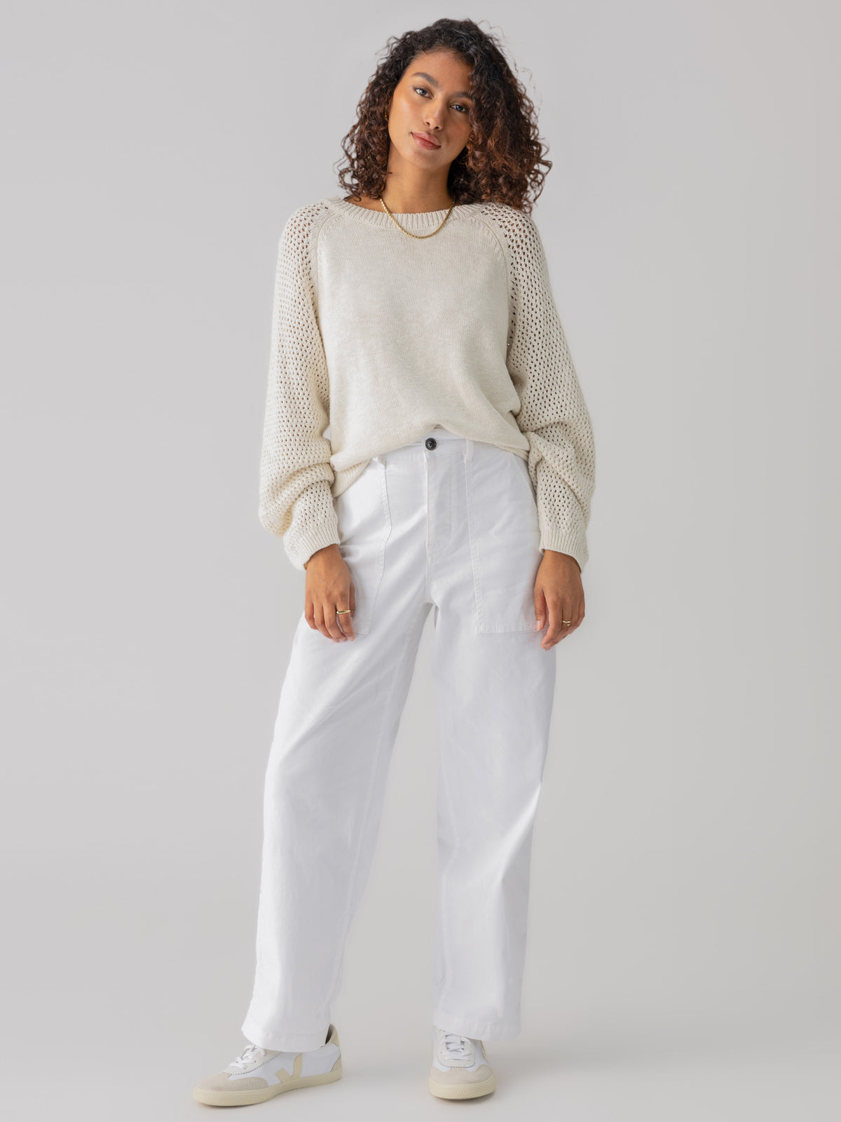 A person with curly hair is standing against a neutral background, wearing the Perfect Moment Sweater Chalk from Sanctuary Clothing, which features mesh sleeves, and white high-waisted pants. They are also wearing white sneakers and looking forward with a relaxed posture.