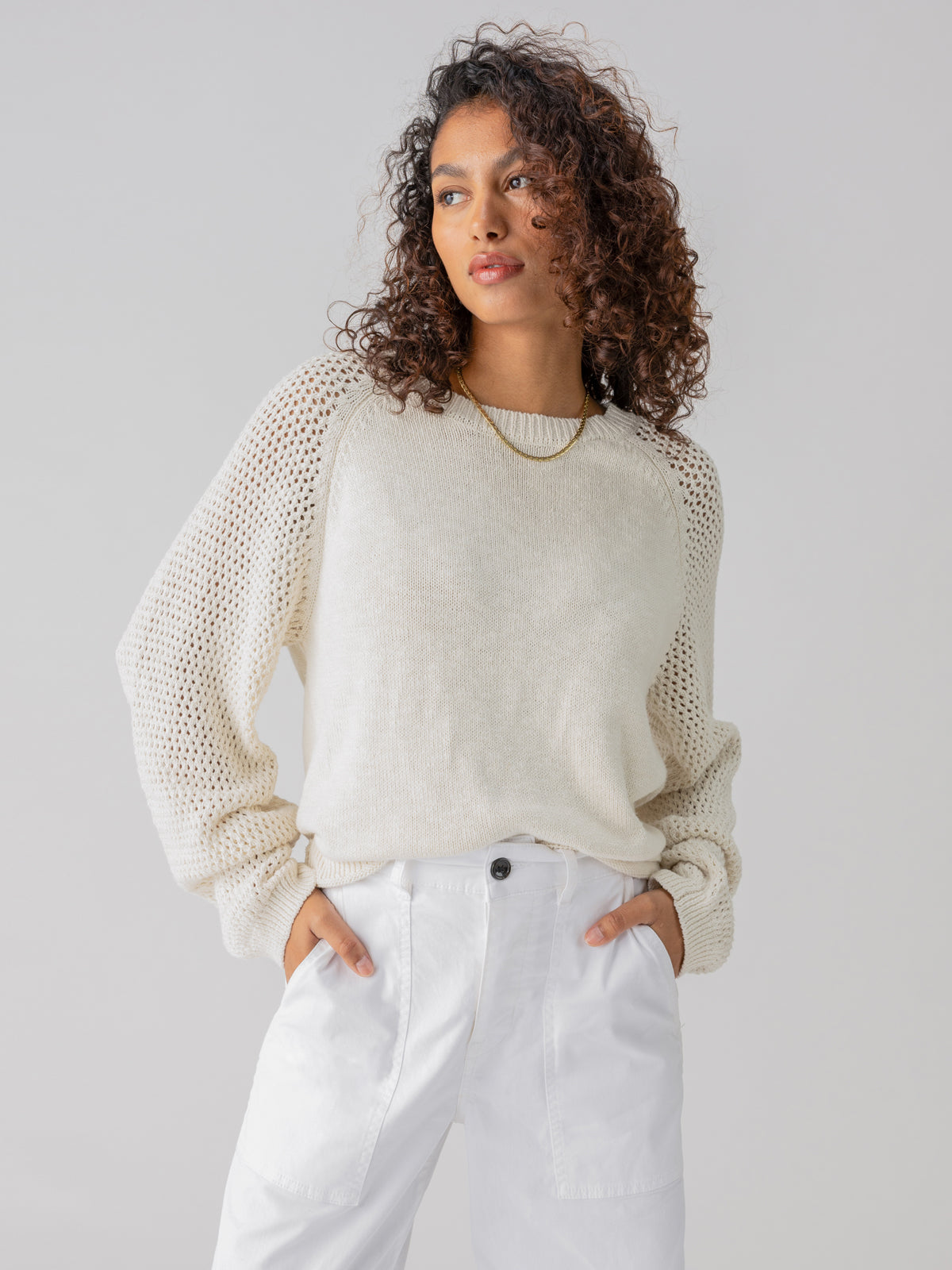 Someone with curly hair is dressed in the Perfect Moment Sweater Chalk by Sanctuary Clothing, featuring mesh sleeves, and paired with white high-waisted pants. Their hands are in their pockets, and they are gazing to the side with a neutral expression. The background is plain and light-colored.