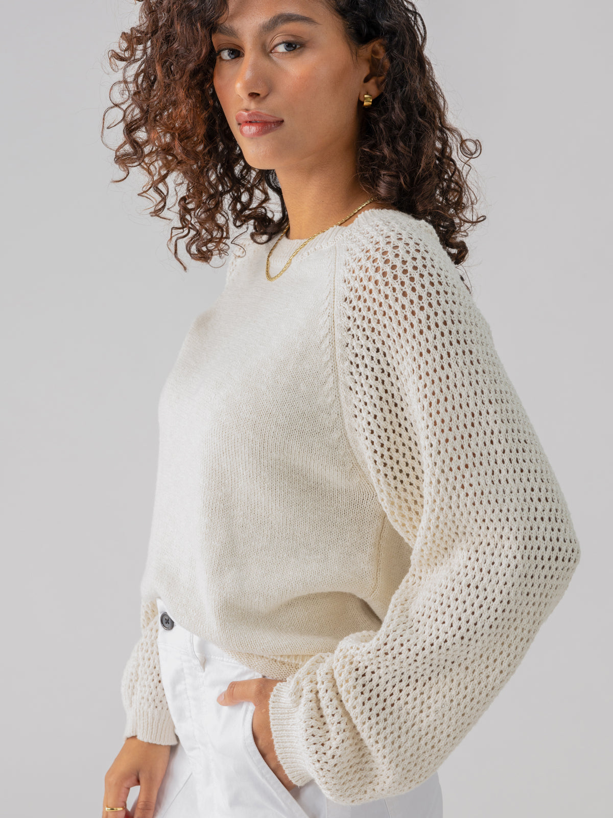 A person with curly hair is wearing the Perfect Moment Sweater Chalk by Sanctuary Clothing, featuring elegant mesh sleeves, paired with white pants. They have a confident expression and are accessorized with a gold necklace and earrings. The background is plain light gray.