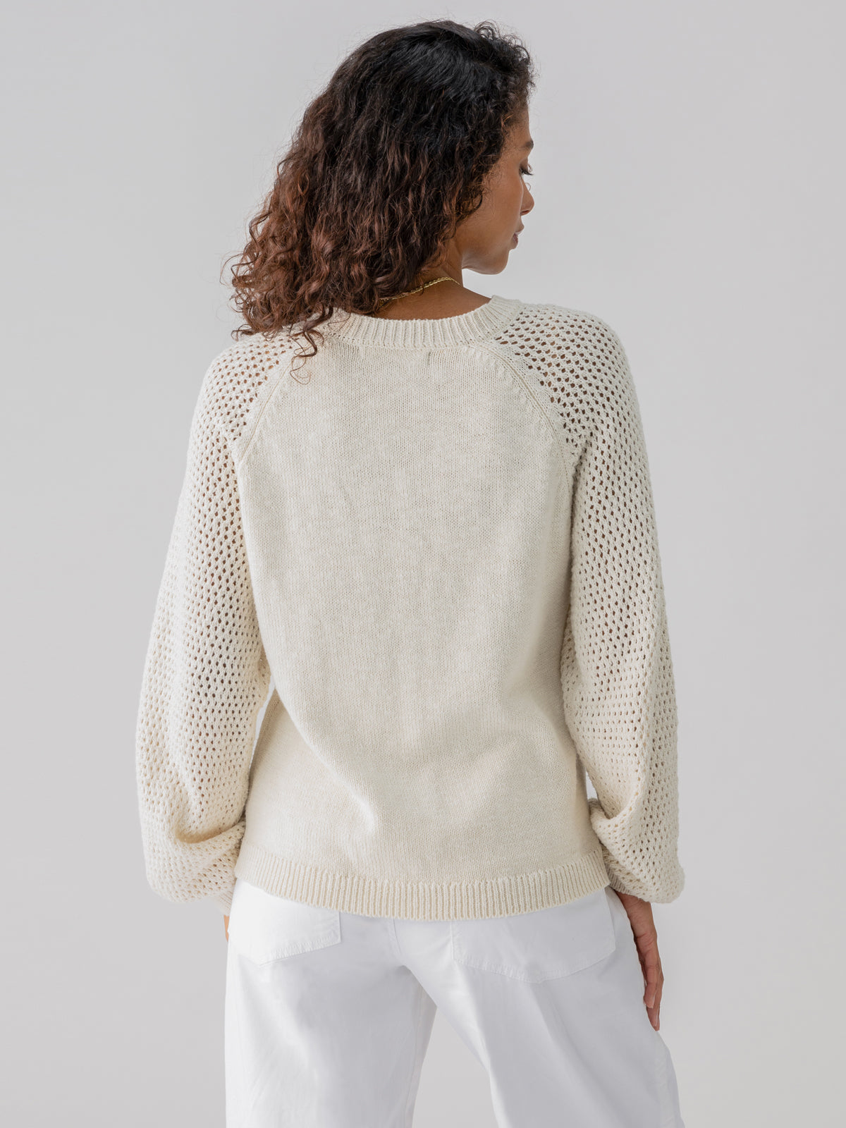 A woman with curly hair stands facing away from the camera, wearing the Perfect Moment Sweater Chalk by Sanctuary Clothing, which features a light beige knitted design with a solid back and mesh-style sleeves, paired with white pants. The background is plain and light-colored.