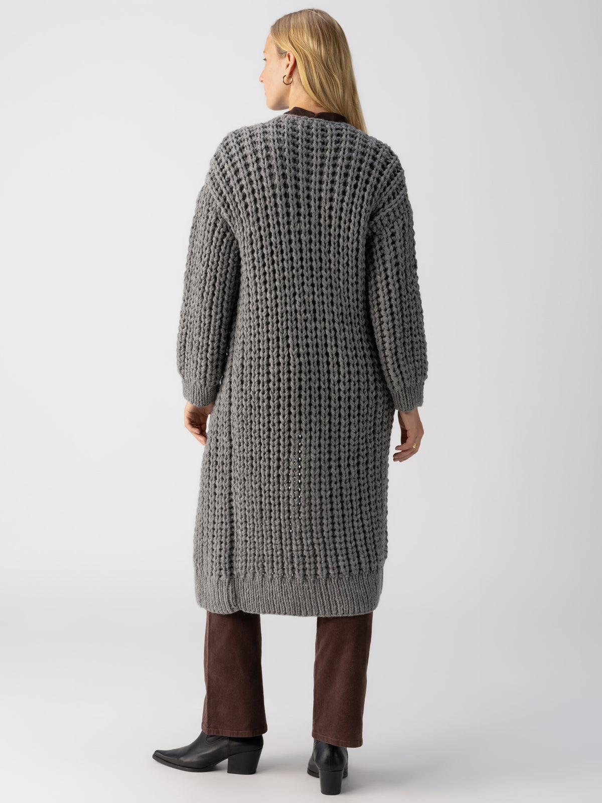 A person with long blonde hair is standing with their back to the camera, wearing "The Ultimate Cardi" in Heather Ash by Sanctuary Clothing. They are also wearing dark brown pants and black ankle boots, in a well-lit indoor setting against a plain background.