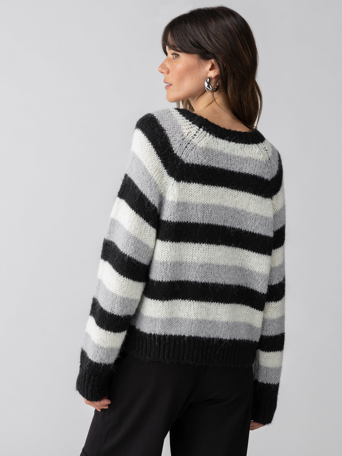 A woman with long dark hair is standing against a neutral background, facing away from the camera. She is wearing the Picture This Stripe Sweater Ash Multi Stripe by Sanctuary Clothing, which features fluffy long sleeves and horizontal black, gray, and white stripes. She has paired the sweater with black pants and hoop earrings while looking over her shoulder.