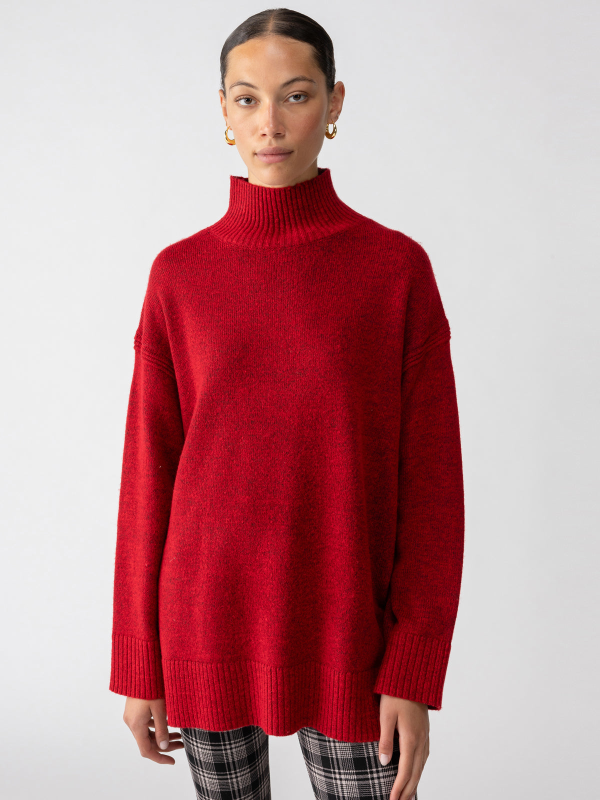 A person dressed in the Perfect Tunic Mars Red from Sanctuary Clothing and plaid pants stands against a plain white background. Their hair is pulled back, and they are adorned with hoop earrings.