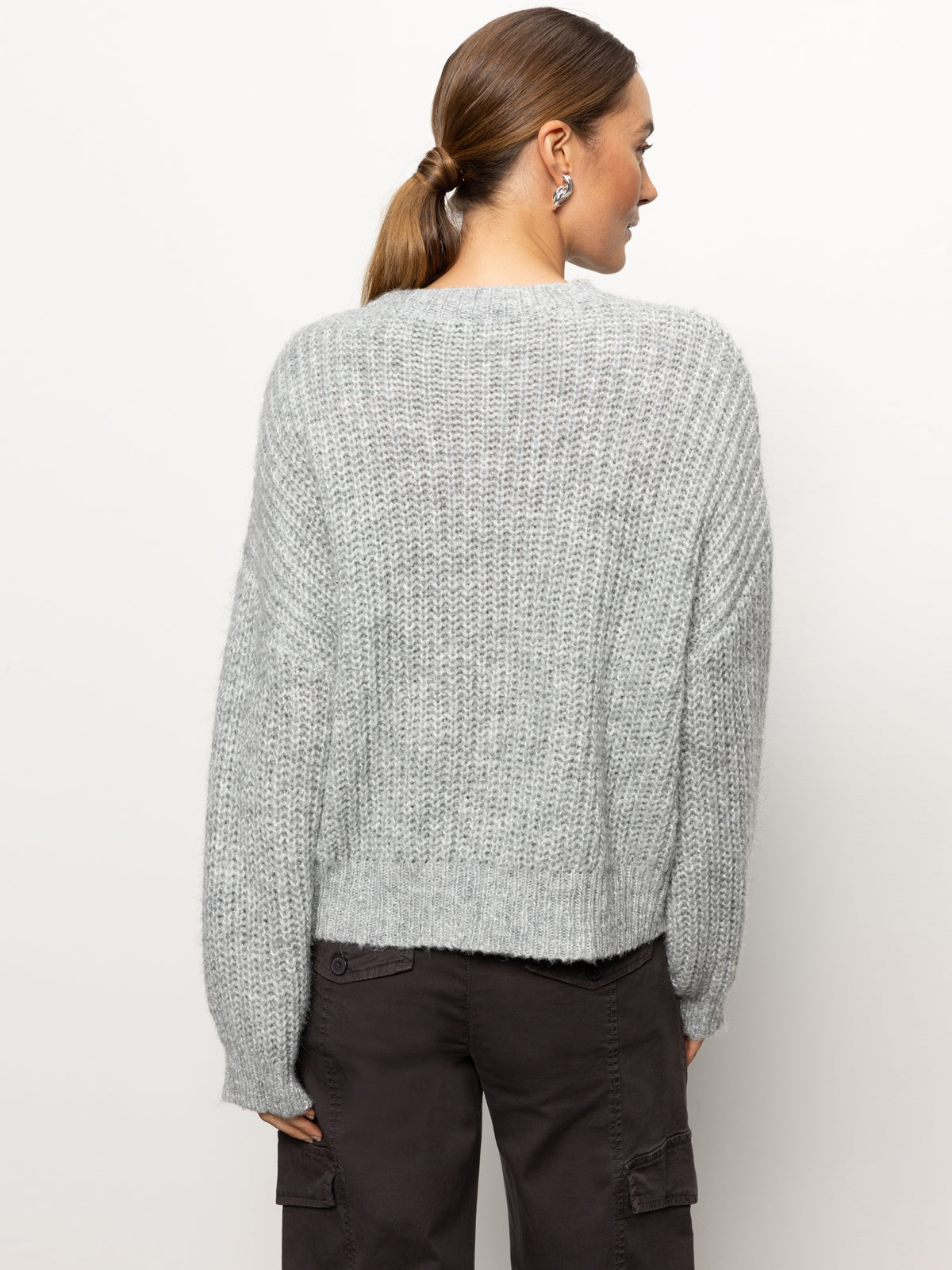 A woman with brown hair tied in a ponytail is wearing the Fuzzy Volume Sleeve Sweater in Heather Grey from Sanctuary Clothing along with dark cargo pants. She is facing away from the camera against a plain white background.