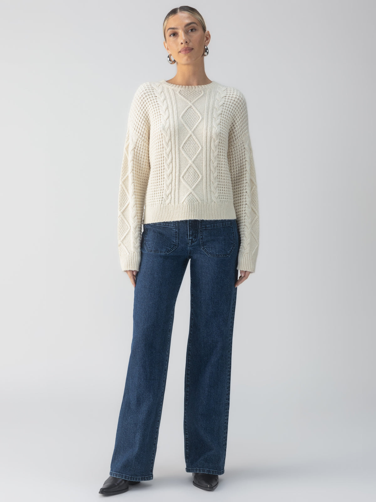 A person stands against a neutral background wearing the Tie Back Cable Sweater in Frosted Almond from Sanctuary Clothing, paired with dark blue jeans. They have their hair pulled back and are wearing black shoes, striking a relaxed and casual pose.