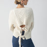 A person with their hair in a bun wears the Tie Back Cable Sweater Frosted Almond by Sanctuary Clothing, which showcases a diamond pattern and cable knit design with an open back and tie detail. They pair it with high-waisted blue jeans and are turned slightly to the side.