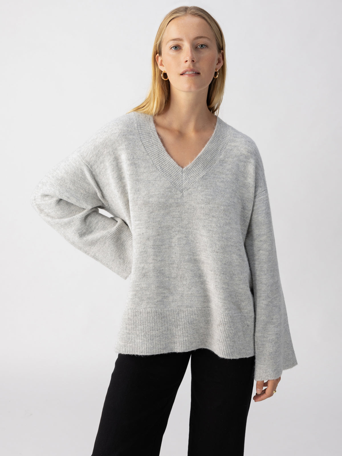 A person with long, blonde hair poses against a plain background, wearing an oversized Sanctuary Clothing Street Chic Sweater in Heather Grey with wide sleeves and black pants. They have a relaxed and confident expression, with one hand resting on their hip.