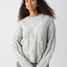 A person with curly hair is wearing a soft, heather grey Cable Crew sweater from Sanctuary Clothing along with grey pants. They are standing with their hands slightly in their pockets against a plain white background. They have a neutral expression on their face.