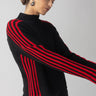 A person dressed in the Sanctuary Clothing Sporty Stripe Mock Sweater Black Multi, featuring red striped sleeves, pairs it with black pants and large hoop earrings. They stand against a gray background, gazing downward with one arm slightly extended.