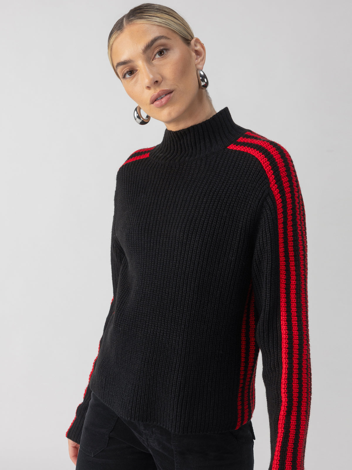 A person wearing the Sanctuary Clothing Sporty Stripe Mock Sweater in Black Multi stands against a plain background. They have blonde hair tied back and are accessorized with large hoop earrings.