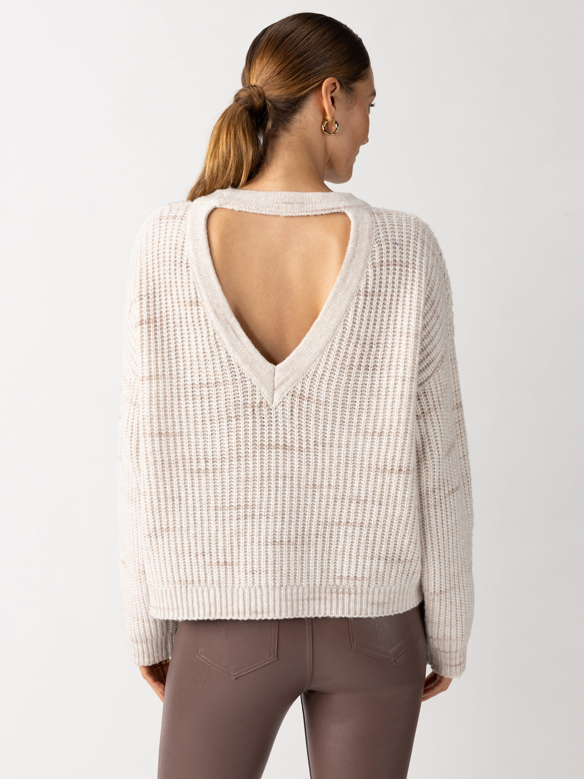 A person with long hair in a ponytail is shown from the back wearing the Cozy V Back Sweater Coco Smoke Multi by Sanctuary Clothing, featuring a large V-shaped open back, paired with brown pants. The background is plain and light gray.