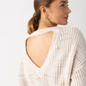 A person with hair pulled back into a low bun is wearing the Cozy V Back Sweater in Coco Smoke Multi by Sanctuary Clothing and gold hoop earrings, facing slightly to the side. The texture of the sweater is ribbed, and the background is neutral.