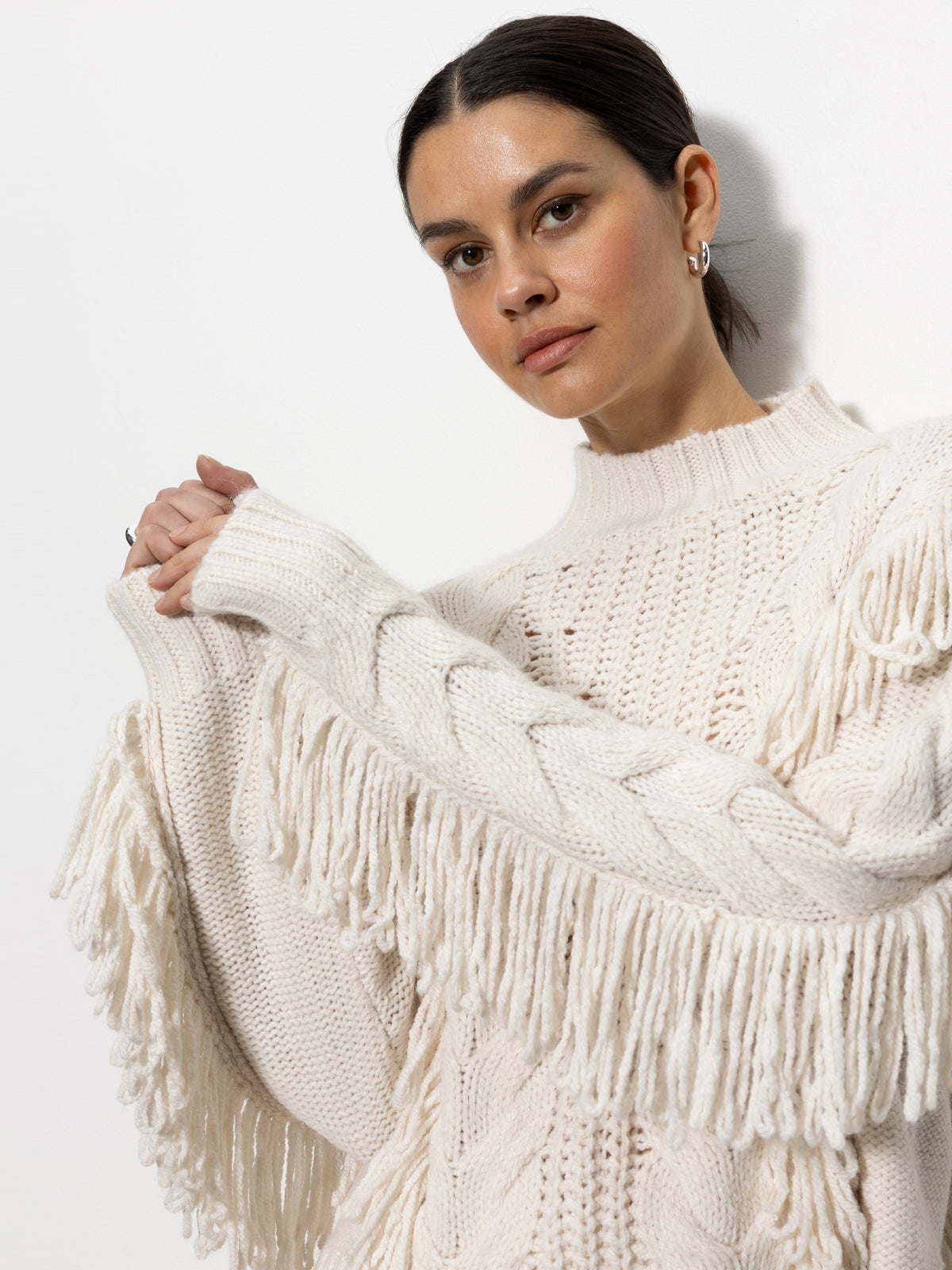 A person wearing Sanctuary Clothing's crafted fringe sweater in chalk stands against a plain background, looking slightly sideways with hands lightly clasped together.