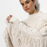 A person wearing Sanctuary Clothing's crafted fringe sweater in chalk stands against a plain background, looking slightly sideways with hands lightly clasped together.