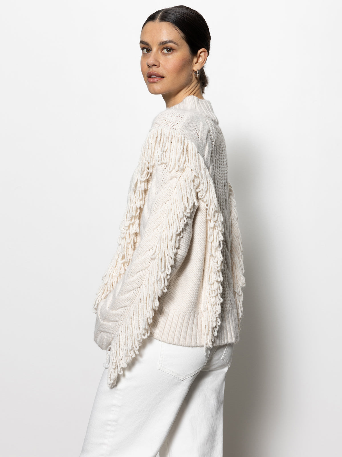 A person models the Sanctuary Clothing crafted fringe sweater in a chalk hue, standing against a plain white background. Their dark hair is pulled back as they pair the sweater with white pants, glancing over their shoulder at the camera.