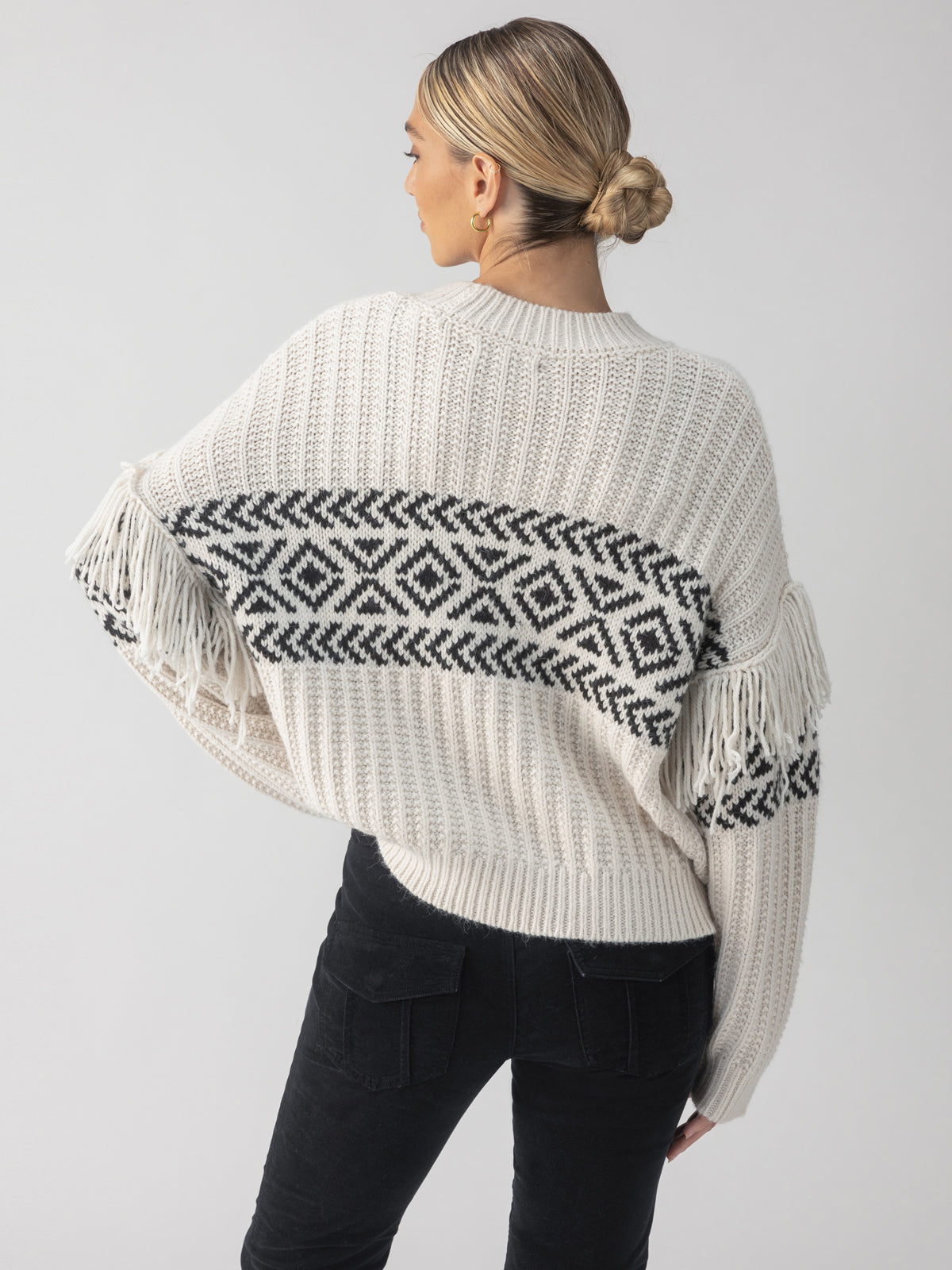 A person with blonde hair styled in a bun is wearing the Artisan Fringe Sweater in Chalk and Black by Sanctuary Clothing, featuring black and white geometric patterns along with fringe details on the sleeves and back. They are facing away against a plain background.