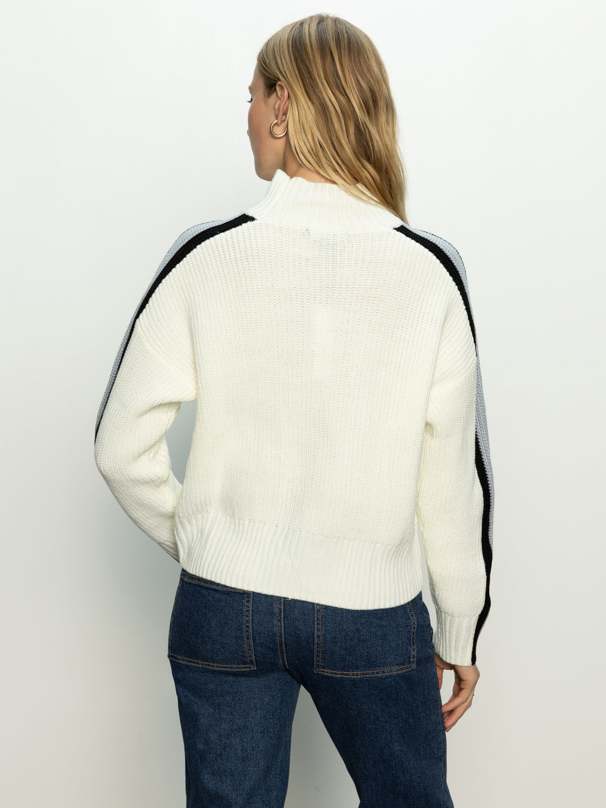 A person with long, light brown hair is wearing the "mock neck stripe sweater" by Sanctuary Clothing, which is pearl blue with black chalk stripes on the sleeves, and blue jeans. They are facing away from the camera against a plain background.