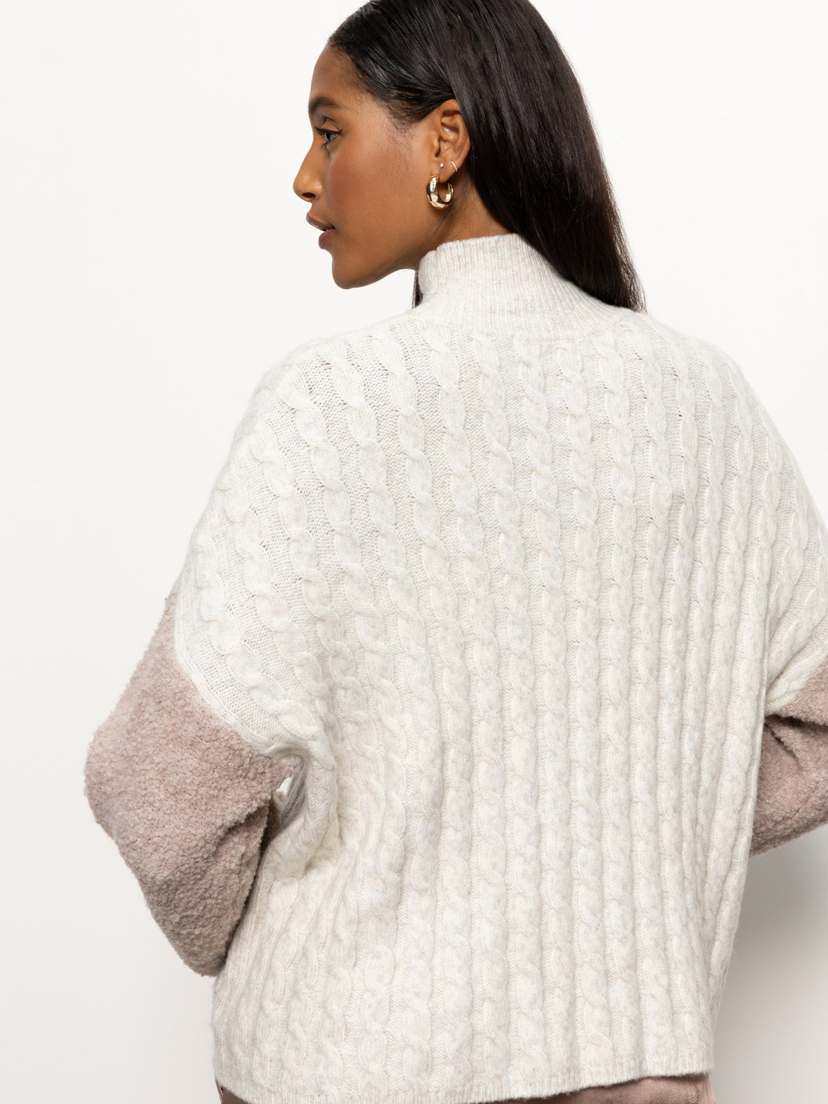 A person facing away is wearing the Sanctuary Clothing's "Boucle Sleeve Sweater" in light heather oatmeal multi, featuring a distinctive cable-knit design and pink sleeves. They have long dark hair and are adorned with gold hoop earrings against a plain white backdrop.