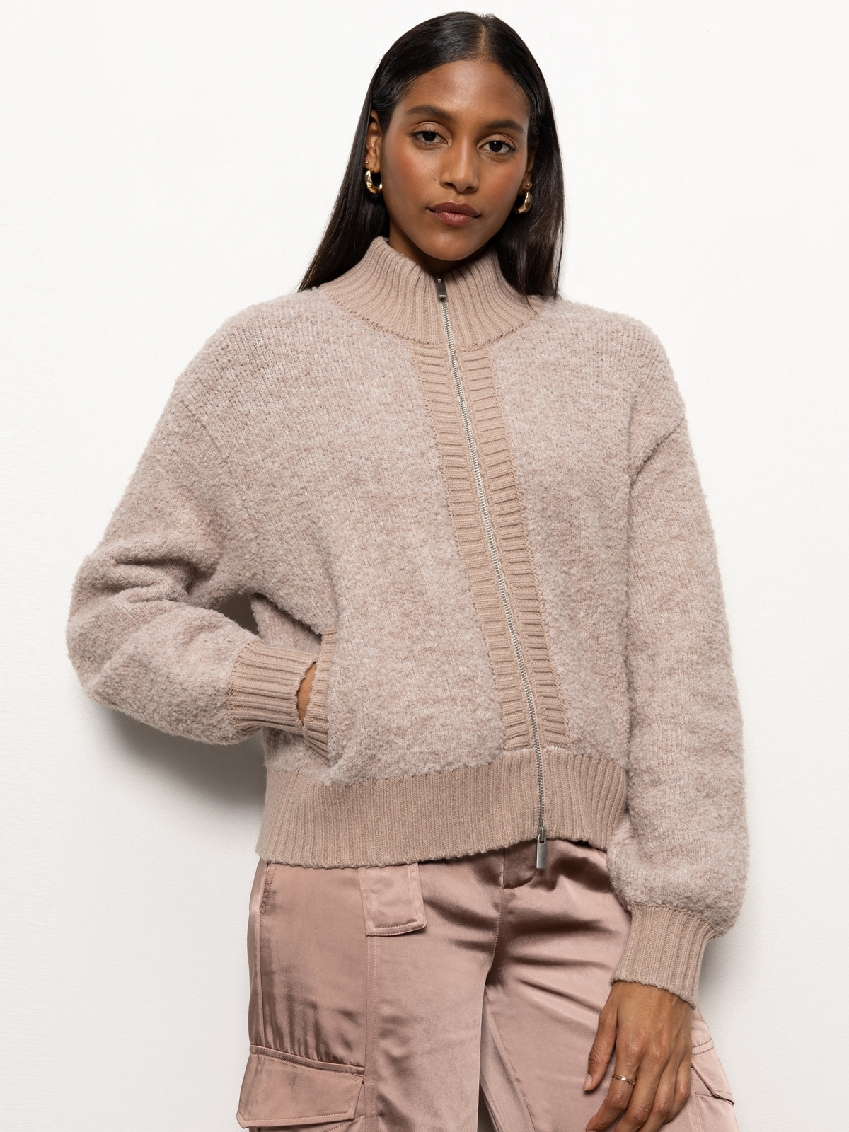 A person wearing the Sanctuary Clothing boucle knit bomber sweater in feather and light brown cargo pants stands against a plain white background. They have one hand in their pocket, are slightly smiling, and feature long dark hair complemented by gold hoop earrings.