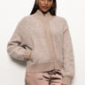 A person wearing the Sanctuary Clothing boucle knit bomber sweater in feather and light brown cargo pants stands against a plain white background. They have one hand in their pocket, are slightly smiling, and feature long dark hair complemented by gold hoop earrings.