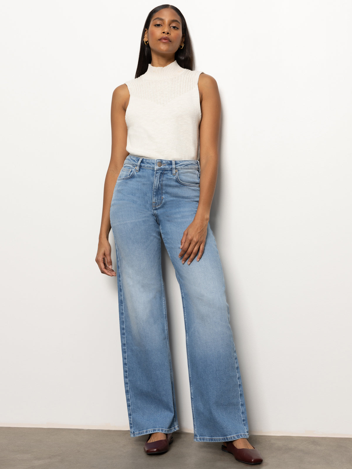 A person stands against a plain wall, dressed in the "rib detail mock tank light oat" by Sanctuary Clothing and light blue wide-leg jeans, paired with brown shoes. They have long dark hair and gold hoop earrings. The pose is relaxed and confident.